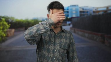 Gözlerini kapayan genç adam, şehir caddesinde, desenli gömlek, modern binalar ve yeşillik ile kentsel dekor giyerek, mahremiyet ve ihtiyatı ifade ederek dur hareketi yapıyor..