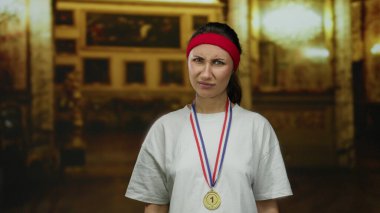 Madalyalı ve bantlı genç bir kadın kilisenin içinde derin düşüncelere dalmış bir ifadeyle, atletik başarıyı manevi ortamla harmanlıyor..