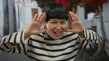 Şehir sokaklarında gülen çiçeklerin altında çizgili süveterle gülümseyen neşeli dış görünüşlü bir kadın şehir ortamında neşe ve doğallığın yakalandığını gösteriyor..