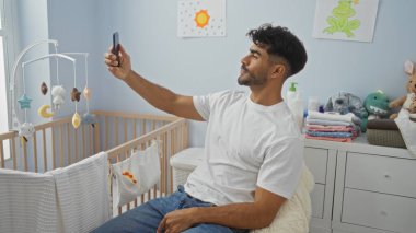 Sakallı genç adam beşikte beşikte selfie çekiyor. Etrafı şen şakrak dekorlar ve renkli sanat eserleriyle çevrili. Ev dekorasyonu sergiliyor..