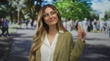 Spor ceket giyen iş kadını, arka planda bulanık kalabalıkla yeşil şehir parkında gökyüzünü işaret ediyor..