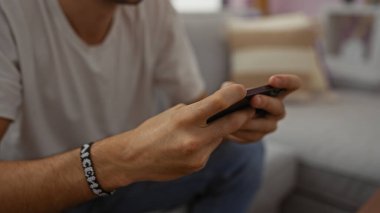 Rahat bir oturma odasında akıllı telefon kullanan genç adam, rahat ambiyansı olan, eller ve teknolojiye odaklanan, modern kapalı alan yaşam tarzını tasvir eden, rahat ve rahat.