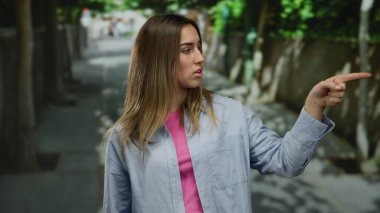 Ağaç kaplı bir caddeyi işaret eden kadın, sıradan bir gömlek giyiyor ve şehir ortamında ciddi görünüyor, odak ve yön anını yakalıyor..