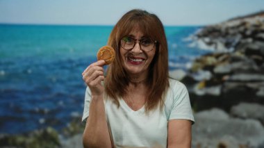 Deniz kenarında gülümseyen İspanyol bir kadın elinde manzaralı bir sahil manzarası ve canlı mavi bir deniz olan bir kurabiye tutuyor..