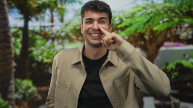 Bir adam, açık havada dikilirken işaret ederek burnunu tutuyor. Gündelik kıyafetler içinde gülümseyen genç bir İspanyol, dost canlısı bir adama benziyor..