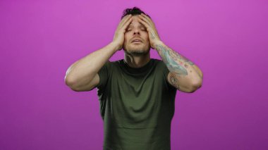 Dövmeleri olan genç İspanyol bir adam pembe arka planda stresli görünüyor, yeşil gömlek giyerken endişeli görünüyor..
