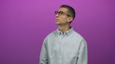 Gözlüklü ve çizgili gömlekli bir adam mor bir duvara bakıyor, arka plandan izole edilmiş, minimalist bir ortamda merak ve bireysellik sergiliyor..