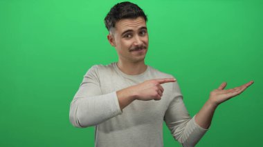 Genç İspanyol bir adam, izole edilmiş yeşil bir arka planda, açık avucunu işaret ederek önerilerde bulunuyor ve dostça bir ifadeyle fikirleri sunuyor..
