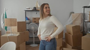 Karton kutular ve mobilyalarla çevrili yeni evdeki kutuları açan bir kadın. Parlak bir oturma odasında kolları ve kalçaları üzerinde durarak enerjik bir şekilde gülümsüyor..