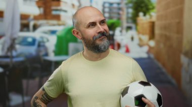 Futbol topu tutan kel bir adam şehir terasında kendine güveniyor, dövmelerini gösteriyor ve sıradan bir şehir manzarasında gülümsüyor..