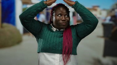 Pembe örgülü, yeşil kazaklı bir kadın ellerini açık havada şehir pazarında açık havada çatı jesti yaparken geniş bir gülümseme sergiliyor..