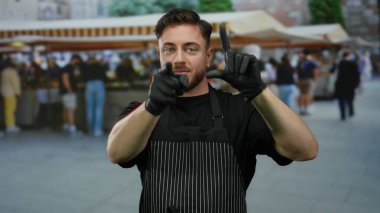 Önlüklü genç sakallı bir adam eldivenli eliyle açık hava pazarında, canlı bir şehir atmosferini yakalayarak ezik bir jest yapıyor..