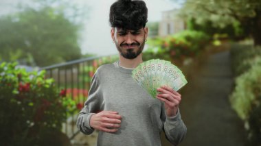 Açık hava parkı ortamında hüzünlü bir ifadeyle Rus ruble banknotları tutan adam, doğal bir arkaplanda mali rahatsızlığı vurguluyor..