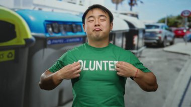Gönüllü gömleği giyen genç adam, şehir sokaklarındaki geri dönüşüm kutularının yanında güvenle duruyor. Çevresel sorumluluğu ve toplumsal bağlılığı teşvik ediyor..