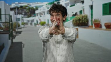 Beyaz tişörtlü bir adam, beyaz binalar ve yeşil pencereli güneşli bir sokakta oyuncak parmaklarıyla silah doğrultuyor..