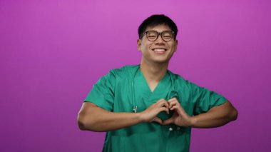 Önlüklü genç adam, elleri pembe izole arka planda kalp şeklinde, gülümsüyor ve gözlük takıyor..