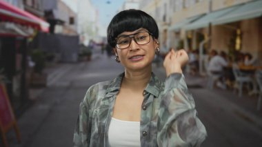 Kalabalık bir şehir caddesinde parmakla gösteren ve gülümseyen bir kadın elleri olan bir kalp yaratıyor, arka planda insanların yemek yediği bir şehir ortamında neşe sergiliyor..