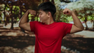 Parkta kaslarını esneten genç Çinli bir adam ağaçlarla çevrili kırmızı gömlekli ve güneş ışığıyla çevrili barışçıl doğal ortamda güç ve kendine güven gösteriyor..