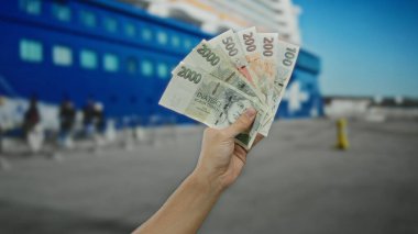 Arka planda mavi bir tekne olan bir sahil limanında Czech Koruna banknotları tutan bir adam açık hava alım satım sahnesini çekiyor..