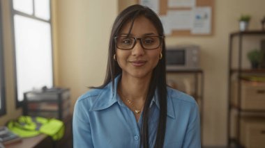 Ofisteki gözlüklü, uzun saçlı ve mavi gömlekli genç bir kadın, kapalı alanda gülümsüyor ve raf arkaplanı olan bir iş ortamında olumlu bir tavır sergiliyor..