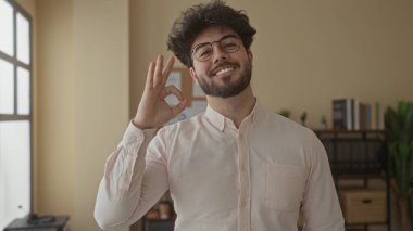 Sakallı ve gözlüklü genç adam ofiste gülümsüyor. Parlak odada bitki ve kitaplarla iyi bir jest yapıyor. İçerde profesyonel ve ulaşılabilir bir atmosfer yaratıyor.