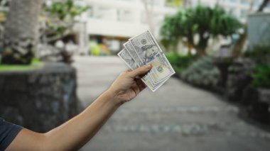 Bir şehir caddesinde Amerikan doları tutan adam, el hareketi ve döviz kuru üzerine odaklanarak şehir yaşam tarzını ve açık havayı ön plana çıkarıyor..