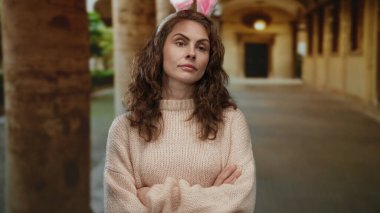 Tavşan kulaklı bir kadın eski bir üniversite koridorunda kollarını kavuşturup akademik ortamın tarihsel atmosferine uygun düşünceli bir ifade sergiliyor..