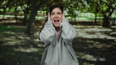 Parkta kulaklarını kapatan, güneşli ağaçların altında günlük giysiler giymiş bir kadın.