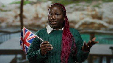 Terasta İngiliz bayrağı tutan kadın yeşil süveter ve kırmızı örgüler giyiyor. Arka planda çok kültürlü kimlikleri simgeliyor. Birlik ve çeşitliliği vurguluyor..
