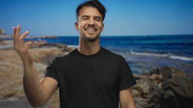 Genç İspanyol bir adam açık mavi bir gökyüzünün altında arka planda okyanusla deniz kıyısında gülümsüyor.