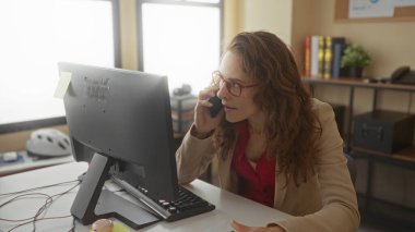 Ofiste telefonla konuşan bir kadın bilgisayarın önünde el kol hareketi yapıyor, iş kıyafeti giyiyor, ofis dekorasyonuyla meşgul ve meşgul bir profesyonel çevreyi canlandırıyor..