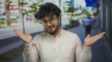 Sokakta duran sakallı ve gözlüklü genç adam bulanık şehir geçmişine karşı açık ellerle şaşkın şaşkın bakıyor..