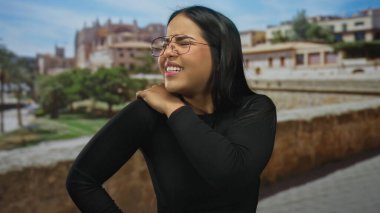 Avrupa 'nın tarihi bir şehrinde omuz ağrısı çeken bir kadın, eski mimari ve yeşilliğin resimsel bir arka planına sahip bir rahatsızlık ve esneklik karışımı sergiliyor..