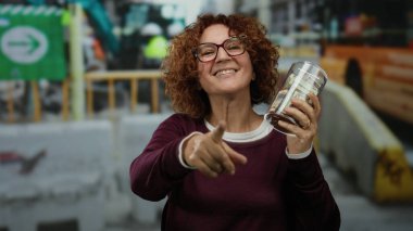 İnşaat alanında elinde dolarlarla gülümseyen bir kadın, açık havada dururken işaret ederek, başarılı bir bağış toplama veya tasarruf çabası sergiliyor..