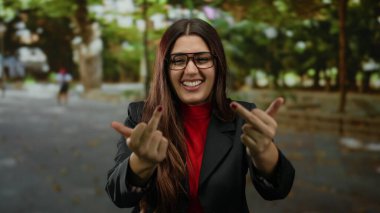 Gözlüklü genç bir kadın, her iki orta parmağıyla bir jest yapıyor. Açık havada bir parkta gülümserken. Etrafı yeşillik ve sonbahar yapraklarıyla çevrili..