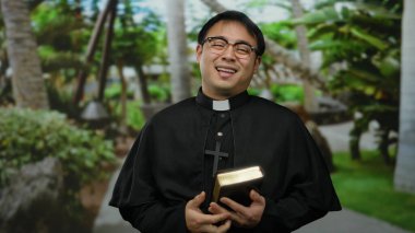 Dini giyinmiş, gözlüklü, gülen bir adam. Elinde bir kitap tutuyor.