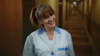 Gülümseyen kıdemli kadın otel koridorunda duruyor, ev ortamında konukseverliği ve profesyonelliği somutlaştırıyor..