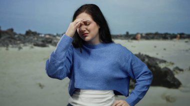 Kayalık bir plajda, deniz ve kumlu çevreyle çevrili bulutlu bir gökyüzünün altında, kapalı gözlerle stresli bir şekilde duran bir kadın..