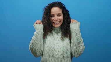 Canlı mavi arka planda, rahat süveter giyen bir kadın stüdyo ortamında izole edilmiş mutluluğu ifade ediyor.