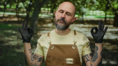 Kel, sakallı ve dövmeli bir adam parkta meditasyon yapıyor. Siyah eldivenler ve sarı tulumlar giyiyor. Gözleri kapalı..