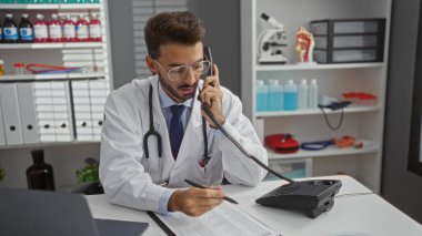 Steteskoplu beyaz önlüklü doktor klinikte hasta kayıtlarını incelerken modern hastane odasının iç dekorasyonunda tıbbi malzemeler arka planda..