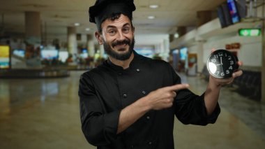 Havaalanı terminalindeki saate işaret eden siyah üniformalı İspanyol şef yoğun seyahat ortamının ortasında zamanın aciliyetini vurguluyor.