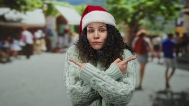 Noel Baba şapkası ve süveter giyen bir kadın ağaçlarla dolu kalabalık bir sokakta neşeli bir şekilde gülümseyerek ve el kol hareketleri yaparak alışverişte arka plandaki insanları bulanıklaştırıyor.