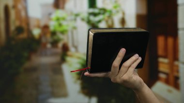 Güneşli bir sokakta elinde kitap tutan bir adam, bulanık yeşil bitkileri olan bir kasabanın huzurlu atmosferini gösteriyor..