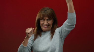 Cesur bir kırmızı arka planda kutlama yapan neşeli bir ifadeyle, İspanyol kıdemli bir kadın mutluluk, enerji ve coşku yayıyor, zafer ve heyecan öneriyor..