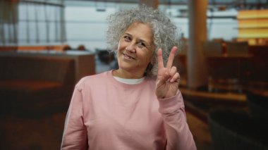 Kapalı bir lokantada huzur tabelasıyla gülümseyen, gri saçları ve günlük kıyafetleri olan bir kadın..