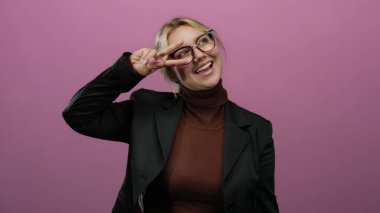 Kameranın önünde barış işareti yapan, sarı saçlı, gülümseyen, pembe arka planda izole edilmiş, gözlüklü ve spor ceketli bir kadın..