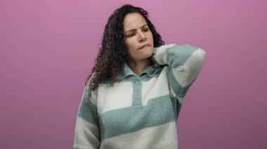 Üzüntülü bir ifadeyle pembe arka planda duran bir kadın, sanki rahatsız olmuş gibi boynunu tutuyor, çizgili bir kazak giyiyor, stresi veya gerginliği vurguluyor.