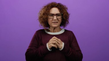 Orta yaşlı, gözlüklü bir İspanyol kadın mor bir duvarın önünde endişeli görünüyor. El hareketleriyle duygularını ifade ediyor..