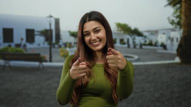 Beyaz binalar ve arka planda yeşil alanlar olan bir sokak ortamında gülümseyen ve şakacı bir şekilde kamerayı işaret eden genç İspanyol kadın..